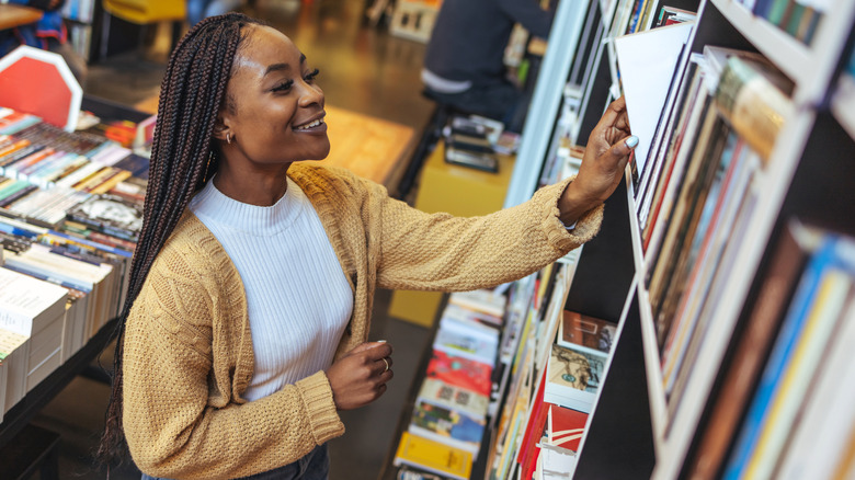 Smiling young woman with braids pulling book from bookstore shelf