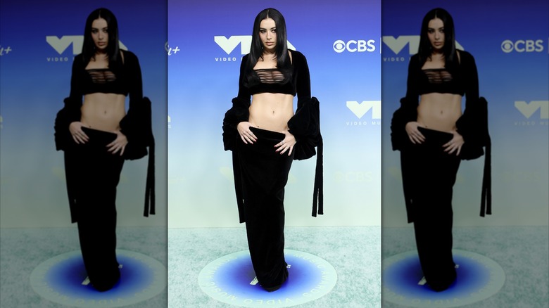 Charli XCX hooking her thumbs into the top of her low-rise skirt while wearing a two-piece ensemble