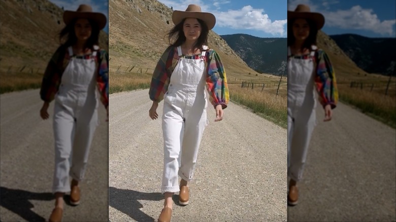 A woman in white overalls with a bright shirt and a cowboy hat on walking outside