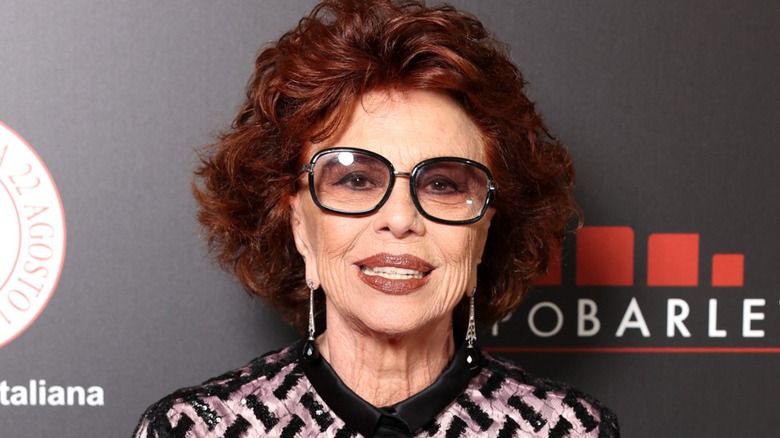 Sophia Loren smiling on the red carpet with voluminous auburn hair and tinted glasses