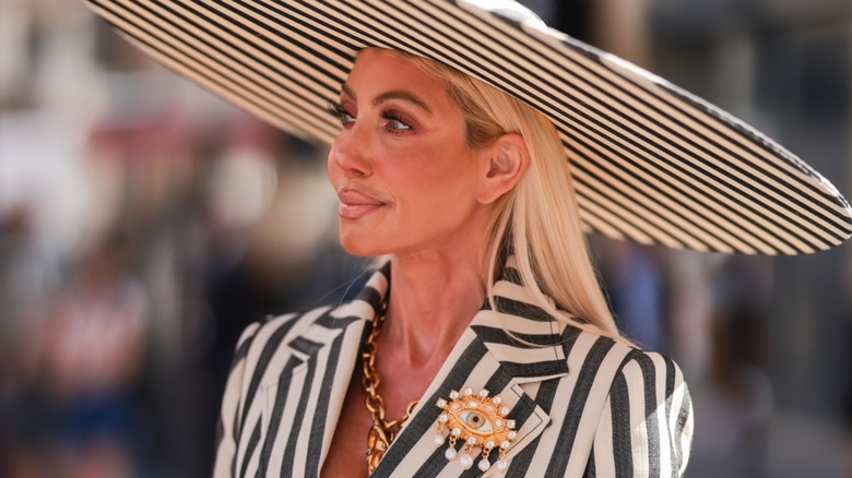 A guest seen wearing a large black and white striped hat, gold large pendant necklace, a gold brooch, a black/white striped blazer jacket and a matching wide leg pants, outside the Schiaparelli show, during Paris Fashion Week - Haute Couture Fall Winter 2022 2023, on July 04, 2022
