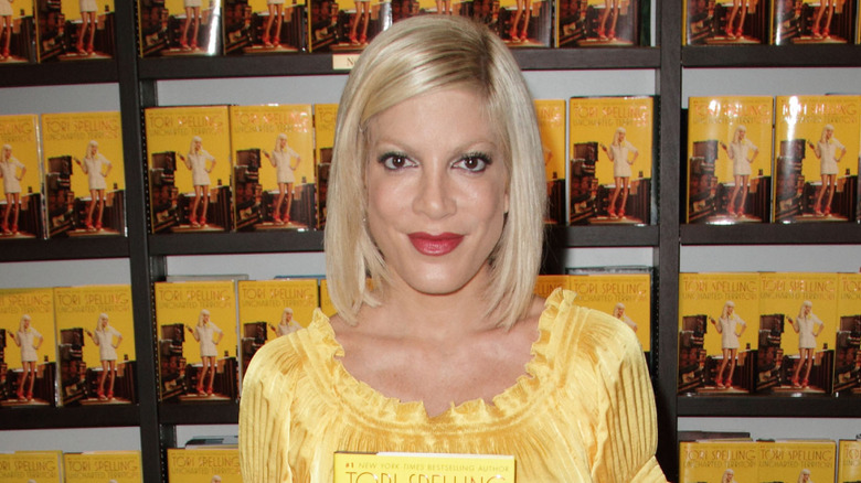 Tori Spelling wearing a yellow shirt, holding her book 