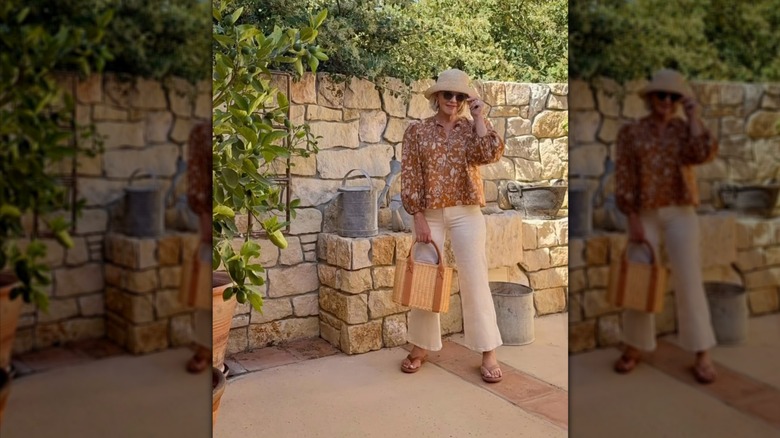 Woman posing on an outdoor patio while wearing a flowy blouse, white pants, and hat