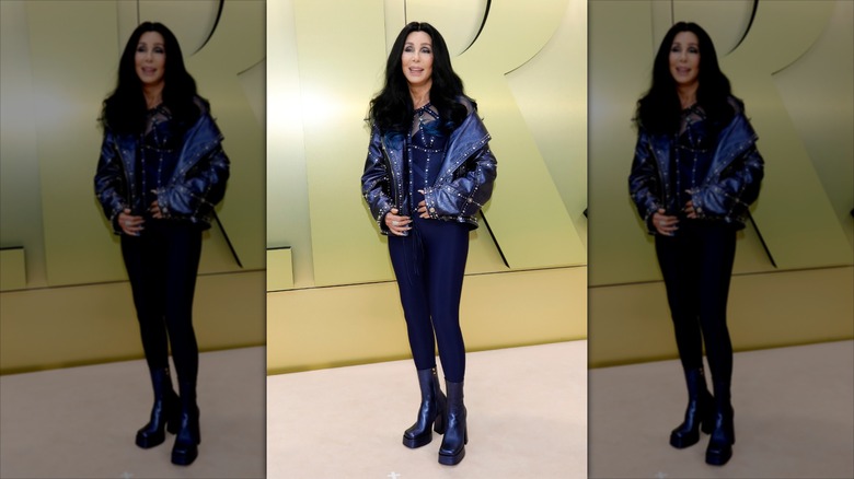 Cher smiles wearing blue disco pants, a leather jacket, and platform boots