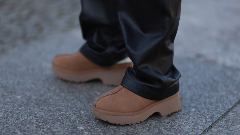 A brown pair of UGG shoes worn with leather pants and pictured against a paved walkway