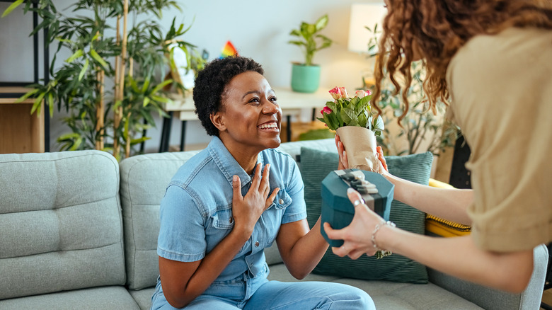 A woman with long hair giving a woman in a denim shirt a box and bouquet of flowers