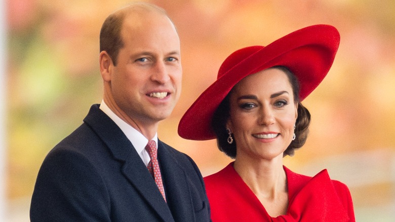 Kate Middleton, in a red dress and hat, smiling alongside Prince William in a suit