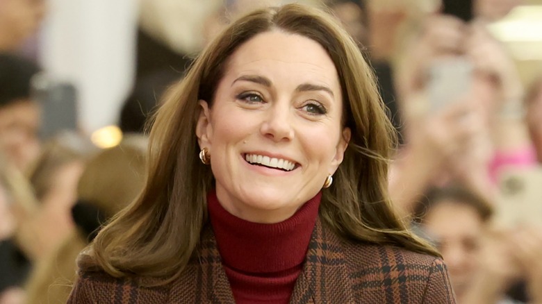 Kate Middleton smiling in a red turtleneck top and plaid jacket
