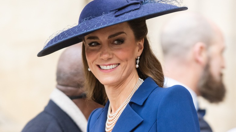Kate Middleton smiling under a large brimmed blue hat while wearing a matching jacket, pearl necklace, and earrings