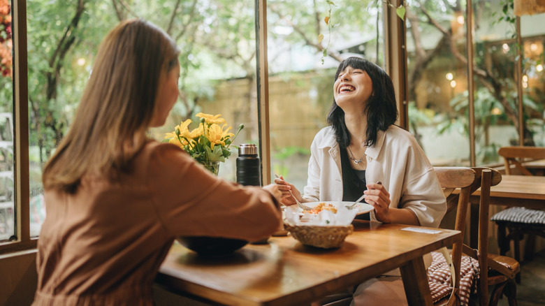 Two women are enjoying a meal together and laughing as they sit near a big window