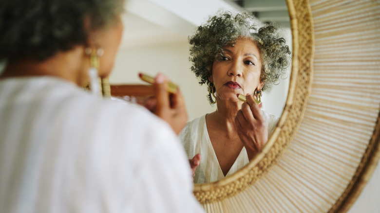 Over-the-shoulder view of an older woman applying lipstick in a mirror