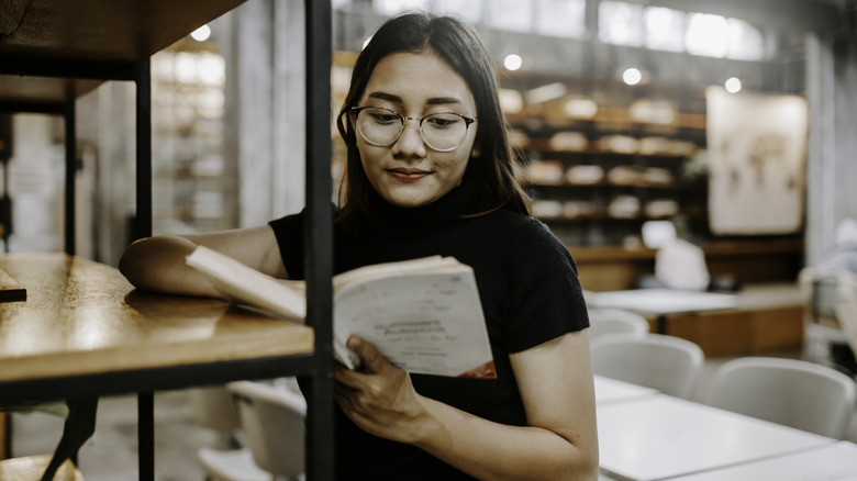 Young, dark-haired woman reading a book while leaning against an industrial bookshelf