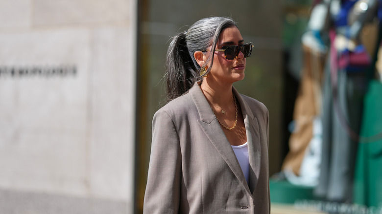 A woman with a gray ponytail and a mushroom toned jacket with sunglasses on outside