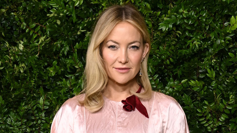 Kate Hudson in a pink dress with a burgundy flower at the neckline standing in front of greenery