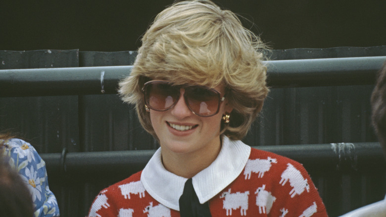 Princess Diana shown from the shoulders up smiling while wearing her 'black-sheep' sweater that was red with white sheep and a black tie.