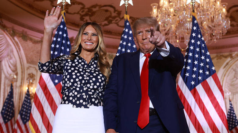 Melania Trump in a white skirt and polka dot top and Donald Trump in a blue suit in front of American flags