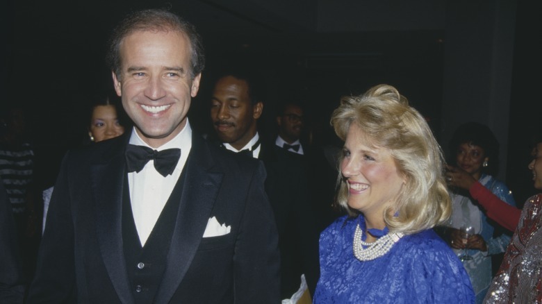 Joe Biden smiles wearing a tuxedo next to Jill Biden wearing a purple dress